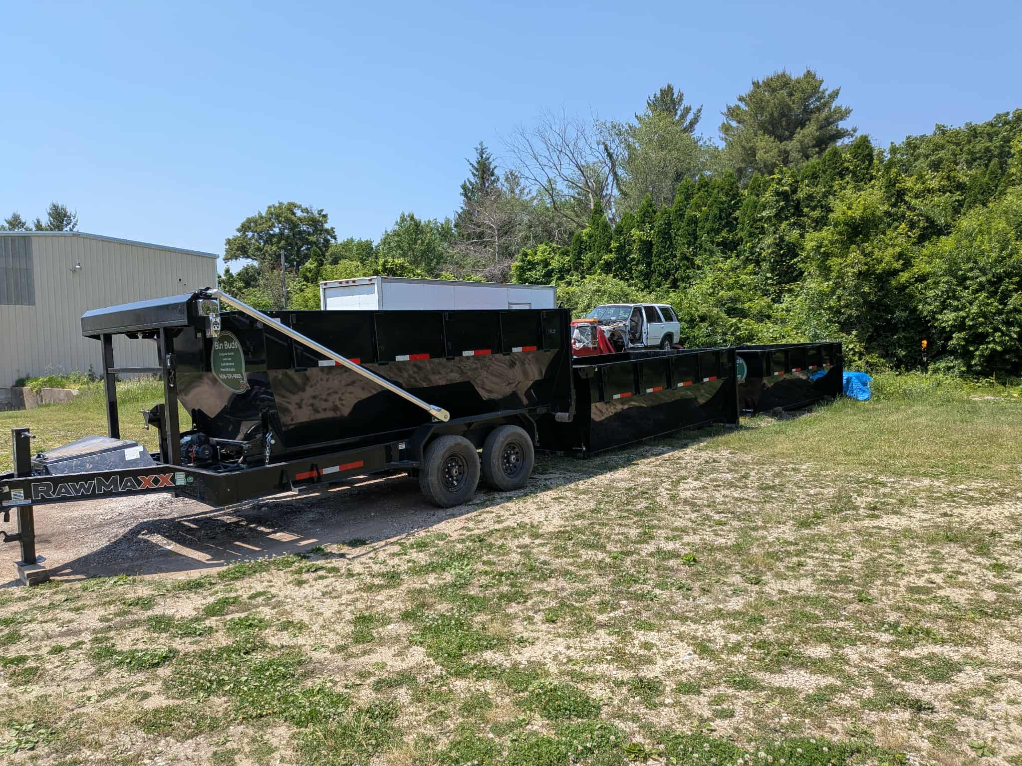 Black trailer and dumpster in grassy area with trees and building