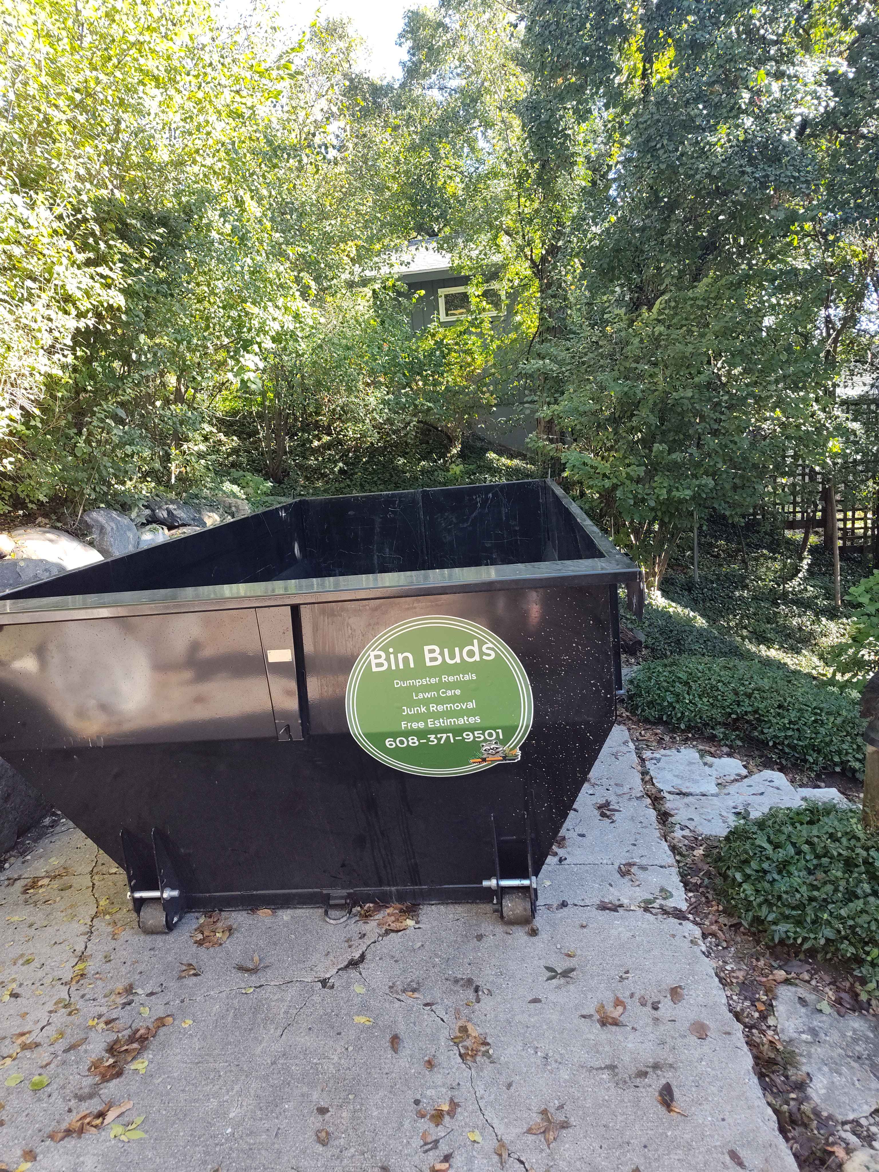 Black dumpster with Bin Buds logo parked on concrete near trees