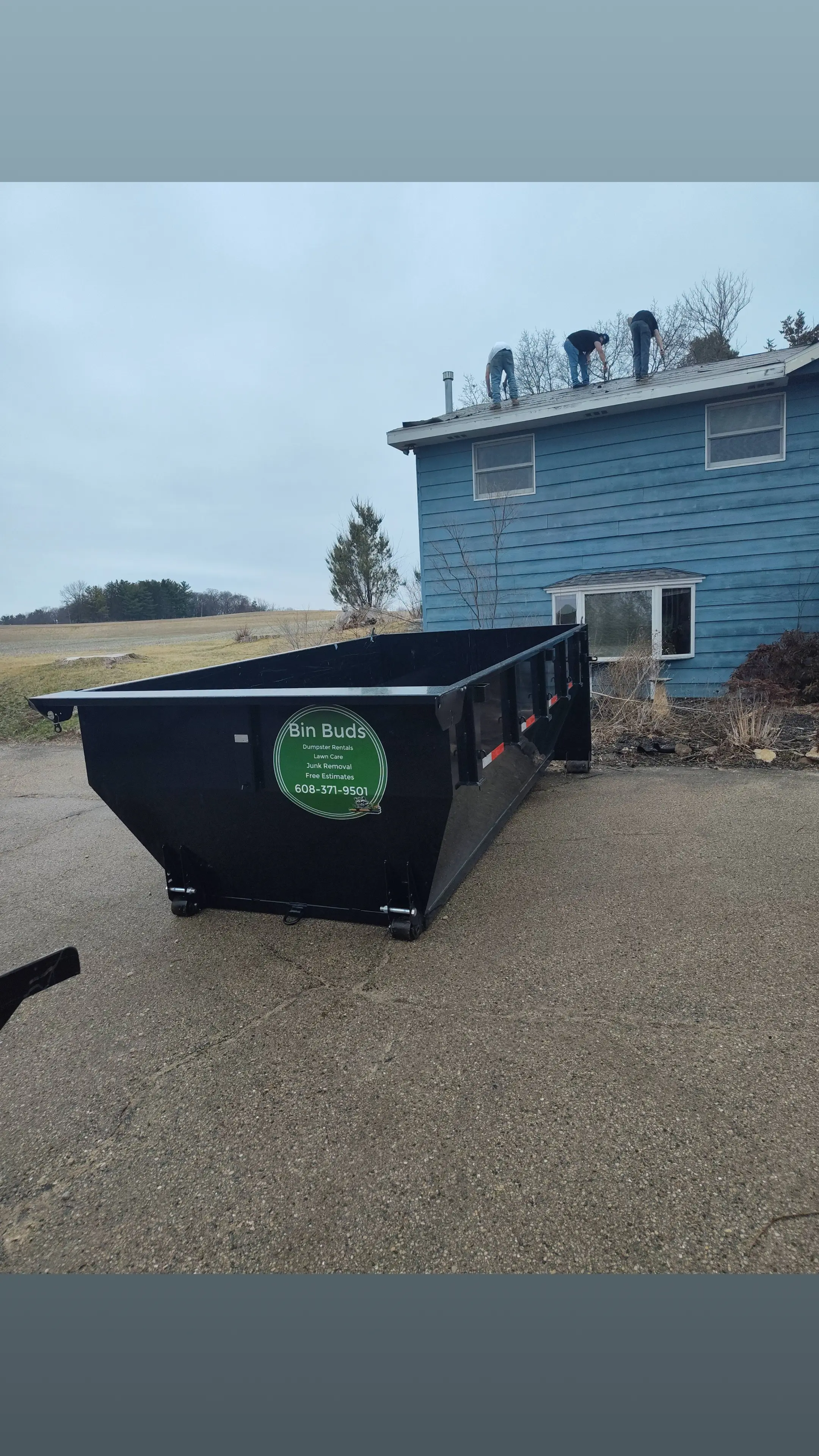 Black dumpster from Bin Buds parked next to blue house with people on roof