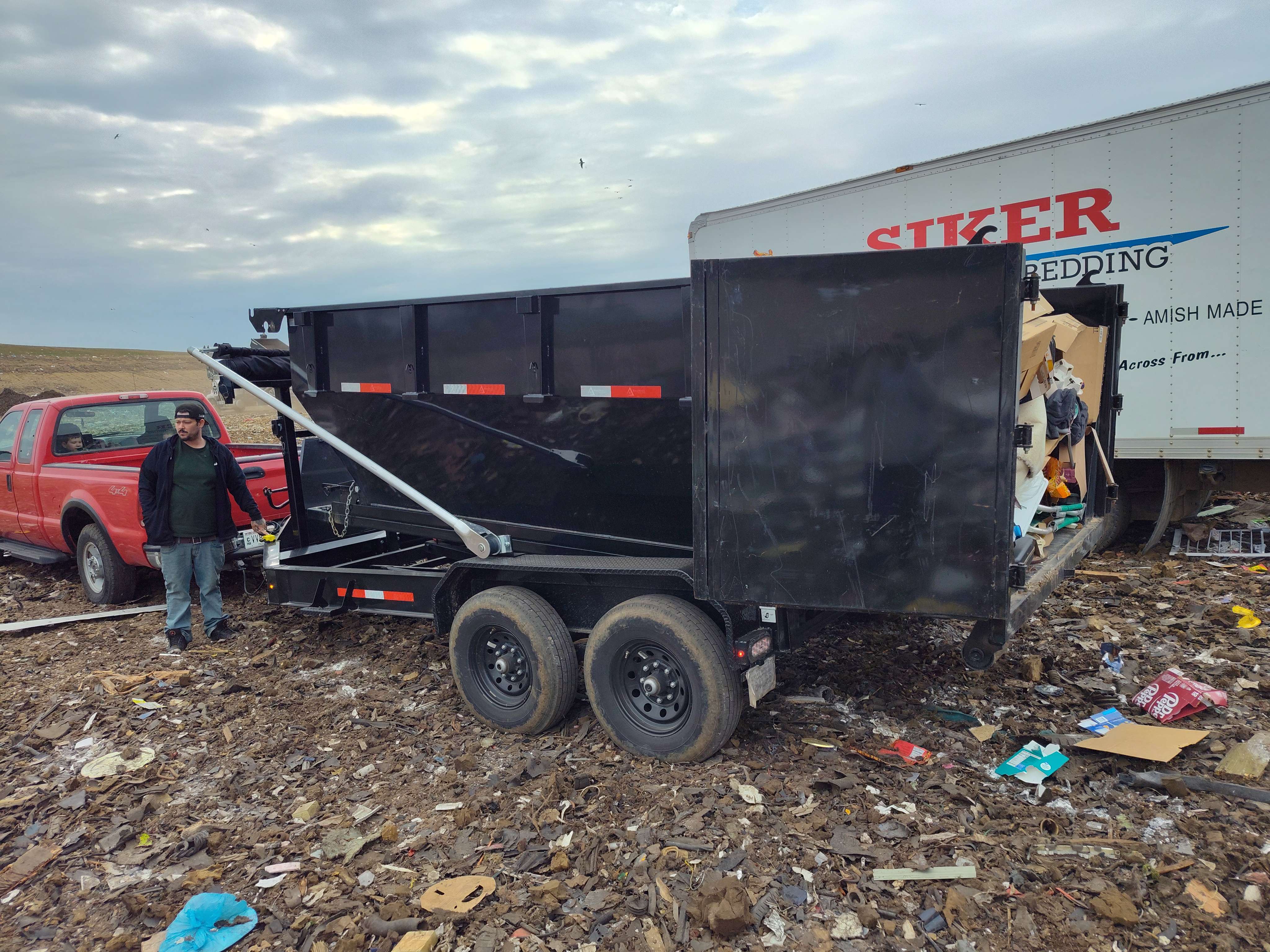 Black dump trailer in littered field with red truck and Stiker trailer
