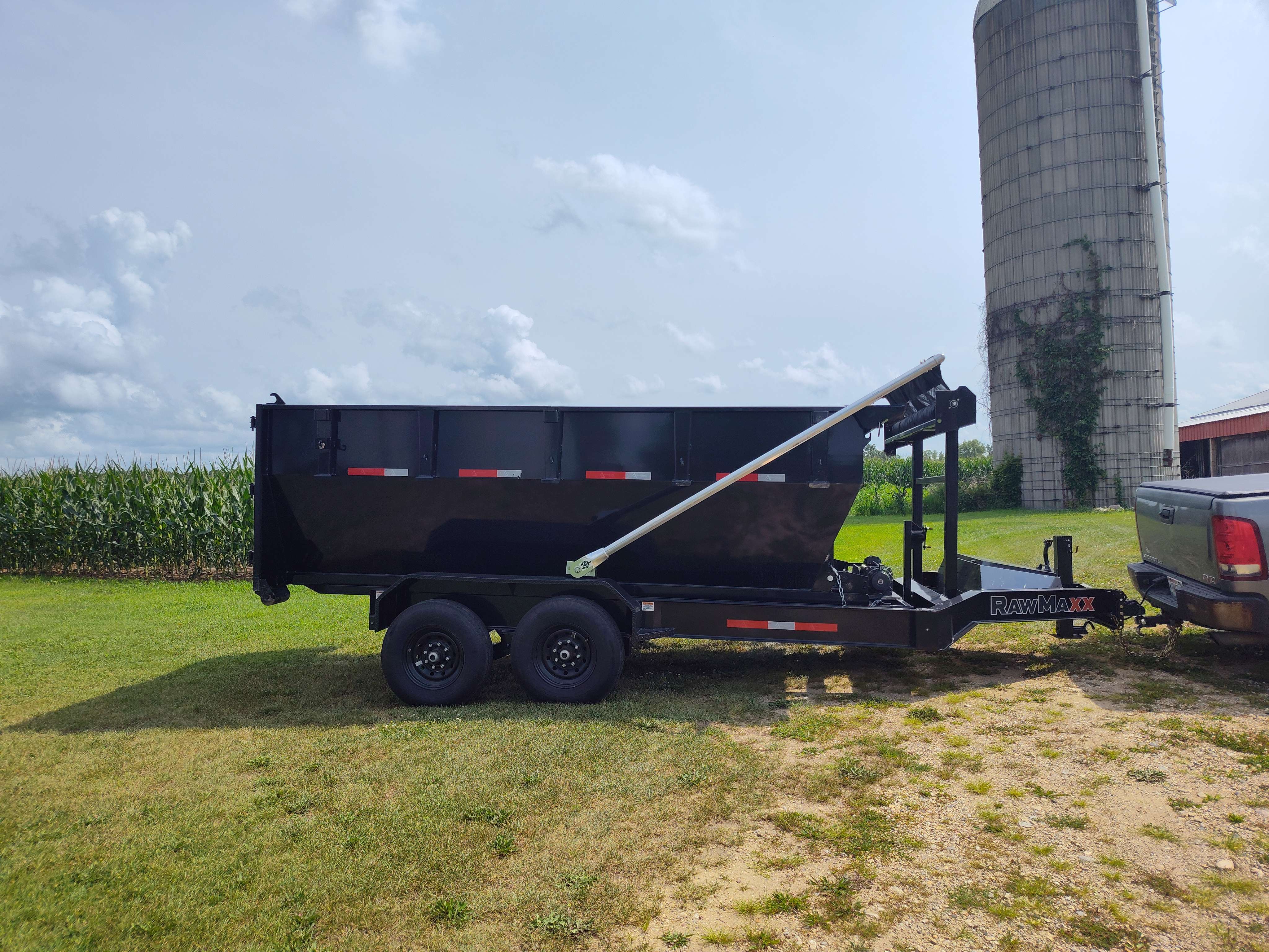 Black RawMaxx dump trailer parked on grass near cornfield and farm silo